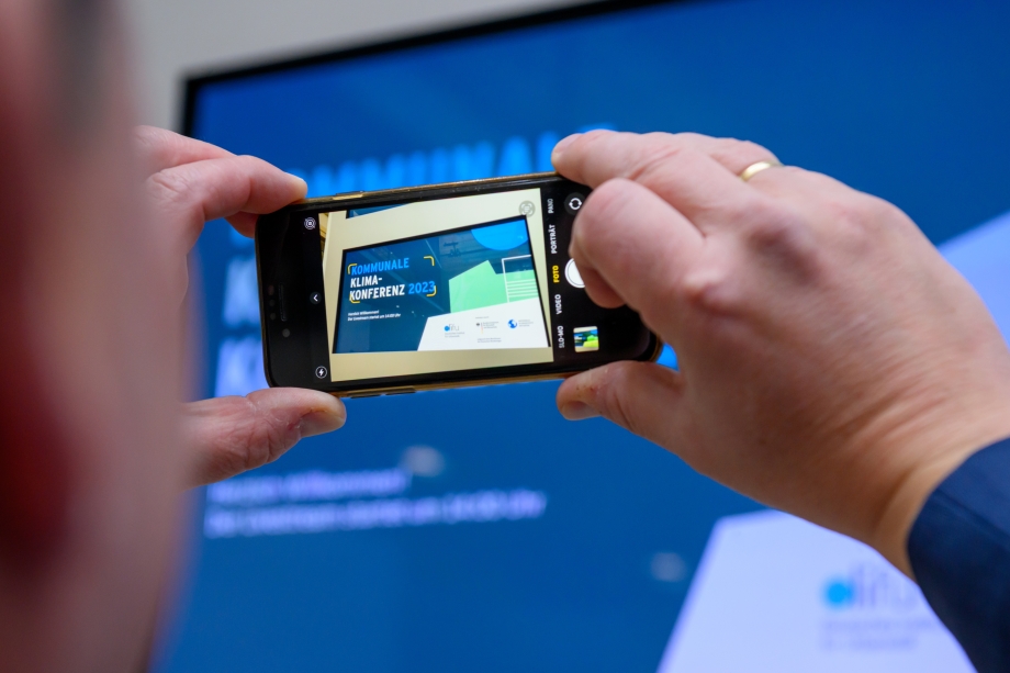 Die Bühne der Kommunalen Klimakonfernz durch den Bildschirm einer Smartphone-Kamera